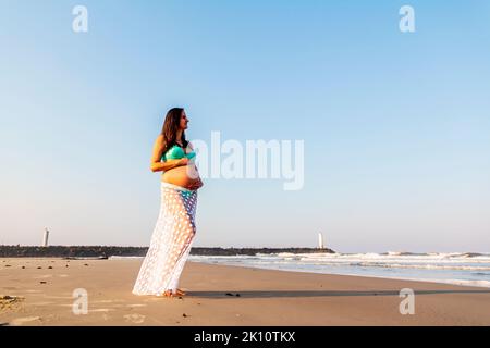 Donna incinta in cima e gonna carezzandole il ventre sulla spiaggia Foto Stock