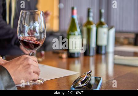 Degustazione di vini a mendoza, Argentina. Mano dell'uomo che tiene un bicchiere di vino rosso con un telefono cellulare e occhiali da sole sul tavolo e bottiglie di vino sfocate Foto Stock