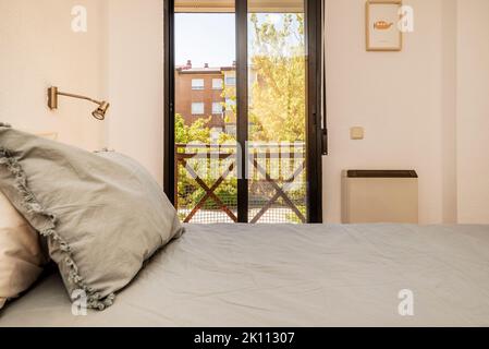 camera da letto con letto matrimoniale con testiera con cuscini grigi, balcone in alluminio marrone con vista sul giardino e dettagli decorativi sulla parete Foto Stock