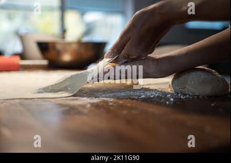 Vista ad angolo basso di una donna che si stende e tira pasta di pasta vegana fatta in casa su un tavolo da pranzo spolverato di farina domestica. Foto Stock