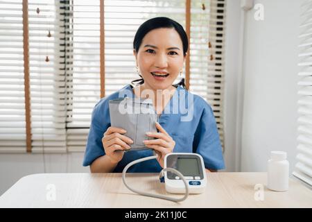 Donna asiatica felice in uniforme blu guardando la fotocamera con sorriso e dimostrando il bracciale del monitor digitale della pressione sanguigna mentre si siede al tavolo e videochiama il paziente Foto Stock