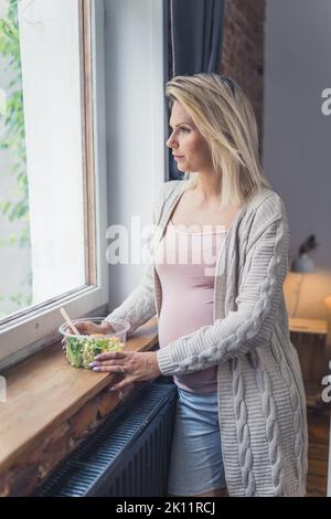 Piano di alimentazione sano durante la gravidanza. Giovane donna incinta caucasica in piedi vicino alla finestra con un nutriente, ricco di vitamine insalata pasto per la crescita di un feto sano. Foto di alta qualità Foto Stock