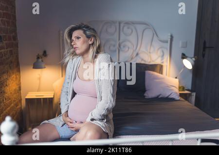 Donna in gravidanza matura che lotta con insonnia, stanchezza e affaticamento durante gli ultimi mesi di gravidanza. Donna seduta su un letto e che sostiene il ventre con le mani. Foto di alta qualità Foto Stock
