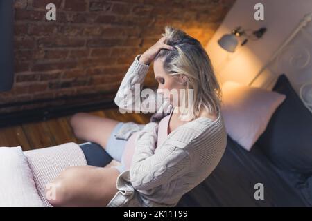 Donna in gravidanza matura che sente dolore allo stomaco e ha un mal di testa durante gli ultimi mesi di gravidanza. Donna preoccupata seduta su un letto e messaging il suo ventre. Foto di alta qualità Foto Stock