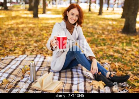 Sorridente signora rosso caucasica millenaria in impermeabile, goditi una tazza di drink, il bel tempo e il tempo libero Foto Stock