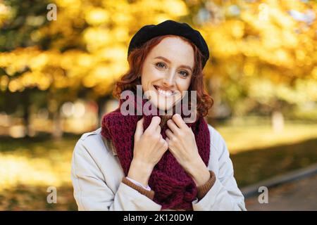 Ritratto di bella donna europea millenario redhead in impermeabile e sciarpa godere del clima caldo nel parco Foto Stock