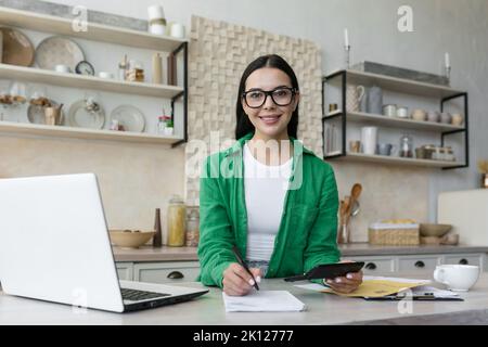 Ritratto di una giovane donna d'affari che lavora a casa on-line remotamente.seduto al tavolo in cucina con un computer portatile, calcolatrice, Documents.in occhiali e una camicia verde, guarda la fotocamera, sorrisi. Foto Stock