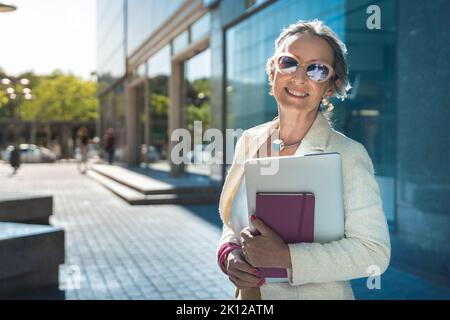 Donna d'affari sicura, concentrata e potente di mezza età che guarda la fotocamera. Bella donna matura sorridente con occhiali da sole, libri e tablet. Foto Stock