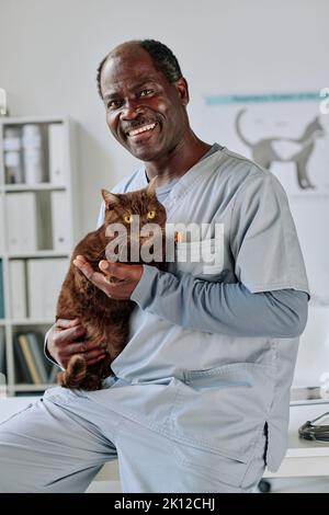 Ritratto di veterinario africano maturo medico in uniforme tenendo gatto domestico e sorridendo alla macchina fotografica mentre lavora in ufficio Foto Stock