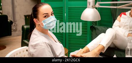 Giovane donna podiatra con una maschera protettiva facendo chiropody e guardando la macchina fotografica nella sua clinica di podiatria Foto Stock