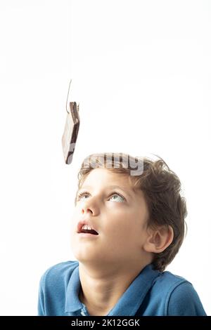 Un bambino guarda a lungo una torta di Spagna appesa ad un gancio. Immagine che mostra il concetto di dipendenza da zucchero e cibi dolci. Sfondo bianco. Foto Stock