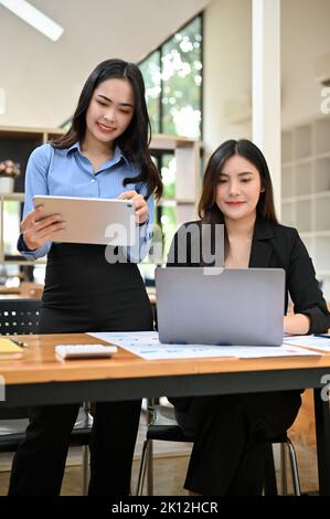 Ritratto, due attraenti donne d'affari asiatiche o dipendenti aziendali che lavorano insieme, utilizzando notebook e tablet in ufficio. Foto Stock