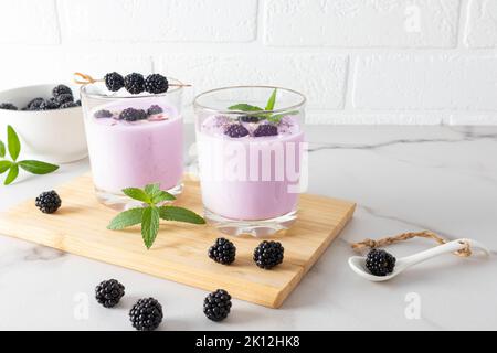 frullato di blackberry in due bicchieri su un asse di legno con more e menta su un tavolo di marmo bianco contro la parete di mattoni. mangiare sano Foto Stock