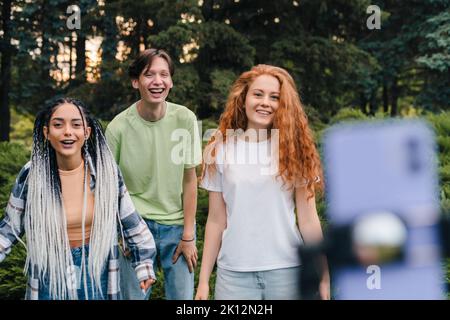 Un gruppo di persone che utilizzano un cavalletto e un telefono ridendo mentre girano video a distanza comunicazione sul telefono cellulare. Registra un video in natura Foto Stock