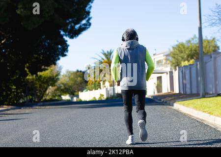 Vista posteriore dell'uomo biraciale anziano in abbigliamento sportivo e cuffie jogging su strada soleggiata Foto Stock