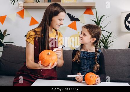 Giovane madre allegra e felice bambina piccola figlia seduto sul divano accogliente pittura piccole zucche arancioni e scherzo, la famiglia si prepara per celebrità Foto Stock