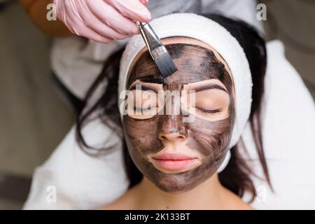 cosmetologo applicazione maschera nera sul viso bella donna indossare guanti neri, donna splendida in spa che hanno le procedure facciali Foto Stock