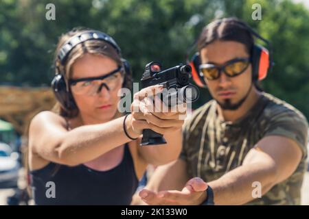 Bell'istruttore maschio in moro T-shirt aiutare una ragazza caucasica a tenere una pistola con una torcia e collimatore correttamente, gamma di tiro al primo piano media. Foto di alta qualità Foto Stock