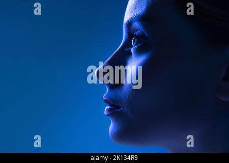 Primo piano vista del profilo di giovane bella ragazza con pelle ben tenuta isolata su sfondo blu scuro in luce al neon. Concetto di arte, moda, stile Foto Stock