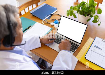Medico di sesso maschile biraciale senior che utilizza il computer portatile e le cuffie per videochiamare, copia spazio sullo schermo Foto Stock