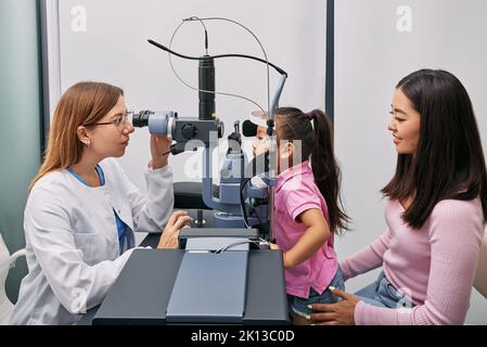 Madre giapponese con la figlia durante la visita all'optometrista. Bambina piccola femmina che ottiene la prova di visione e l'esame dell'occhio in clinica di oftalmologia, lato vi Foto Stock