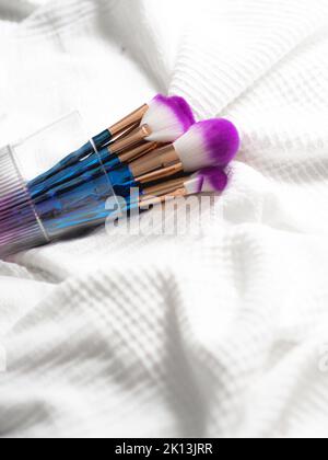Un primo piano di un set colorato di spazzole per il trucco in una tazza di vetro giacente sul lenzuolo Foto Stock