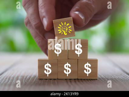 Simbolo del dollaro su blocchi di legno con mano che tiene un blocco di legno con lampadina gialla. Ricchezza e idea concetto Foto Stock