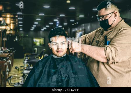 Barbiere con maschera che dona un taglio di capelli al Latino in barbiere Foto Stock