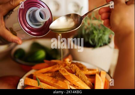 Attenzione selettiva a versare l'olio d'oliva sul cucchiaio e spruzzarlo su spicchi di patate dolci biologiche, mentre si prepara il pranzo Foto Stock