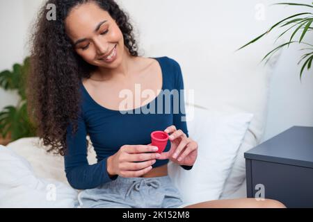 Una bella giovane donna tiene la tazza mestruale mentre si siede sul bordo del letto Foto Stock