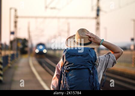 Viaggio in treno. Donna con cappello e zaino in attesa di treno alla stazione ferroviaria Foto Stock