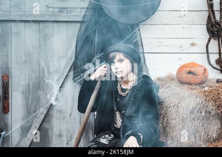 Bella paurosa bambina che celebra Halloween. Terrificante nero, bianco mezza faccia trucco, costume strega, elegante immagine. Divertimento alla festa dei bambini in b Foto Stock