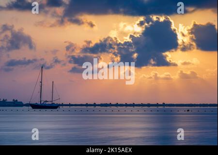 Alba sul Mar Mediterraneo, in una mattinata nuvolosa d'estate. Il sole si riflette sul mare calmo, su cui ci sono alcune barche a vela Foto Stock