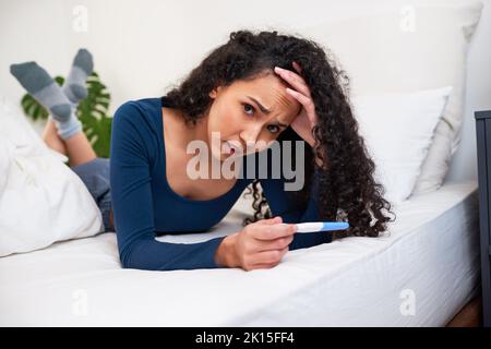 Una giovane donna multietnica si trova a letto guardando i risultati indesiderati del test di gravidanza Foto Stock