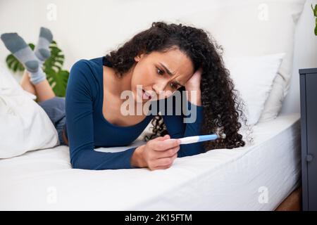 Una giovane donna multietnica si trova a letto guardando i risultati indesiderati del test di gravidanza Foto Stock