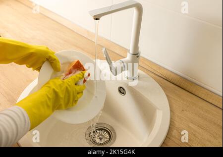 Vista dall'alto delle mani femminili con guanti protettivi lavando le stoviglie sotto l'acqua del rubinetto nel lavandino Foto Stock