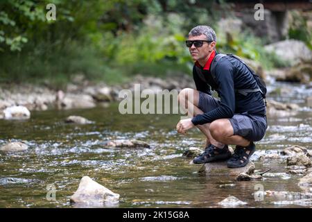 Uomo maturo escursionista accovacciato accanto al fiume di montagna nella foresta Foto Stock