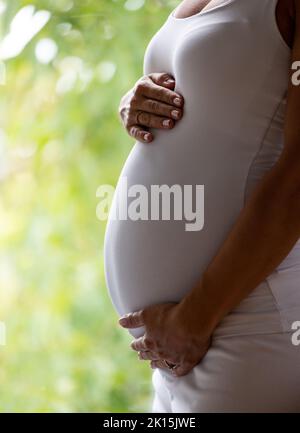 Primo piano del ventre di una donna incinta in una camicia bianca che tiene le mani sullo stomaco. Sfondo verde Foto Stock