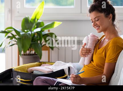 Donna incinta entusiasta valigia di imballaggio per il lavoro e guardando la lista di controllo sul divano a casa Foto Stock