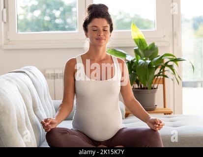 Donna incinta seduta in posizione loto e meditando sul divano in soggiorno Foto Stock