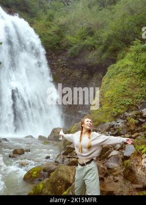 Donna sorridente con una bottiglia d'acqua si alza con le braccia separate vicino ad una grande cascata Foto Stock