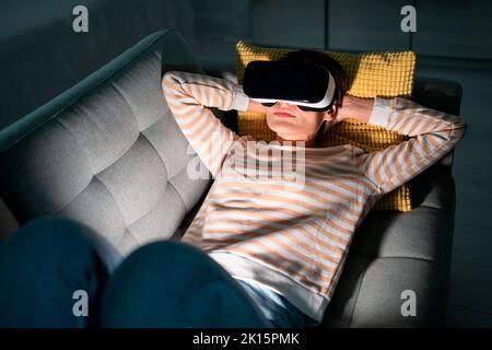 Donna stupita che prova il moderno casco VR high-tech, per vivere l'esperienza 3D a casa riposandosi sul divano Foto Stock