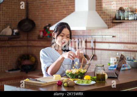 Felice giovane donna asiatica con lunghi capelli scuri in abiti casual sorridendo mentre si è in piedi al tavolo con il computer portatile e gli ingredienti e la preparazione deliziosa Foto Stock