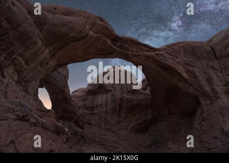 Vista mozzafiato dell'arido parco nazionale degli Arches attraverso la formazione del doppio arco contro la Via Lattea incandescente nel cielo notturno dello Utah, USA Foto Stock