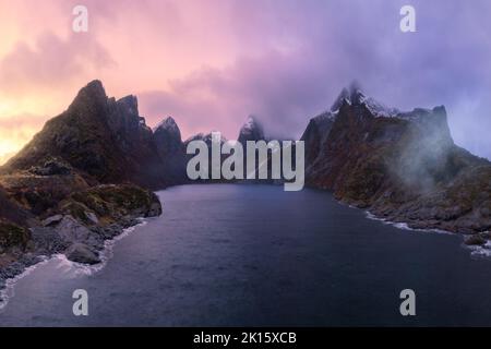 Paesaggio panoramico di montagne rocciose con picchi aguzzi ricoperti di neve vicino al mare calmo contro il cielo tramonto in Norvegia Foto Stock