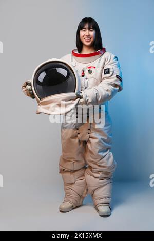 Giovane donna asiatica nella tuta spaziale che tiene il casco e guarda la macchina fotografica sotto la luce blu prima della missione su sfondo grigio Foto Stock