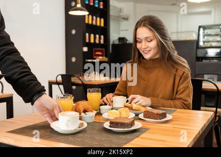 Giovane uomo corto irriconoscibile in uniforme nera prendendo un bicchiere di succo dal vassoio mentre serve cibo per donna sorridente durante la colazione nel ristorante Foto Stock