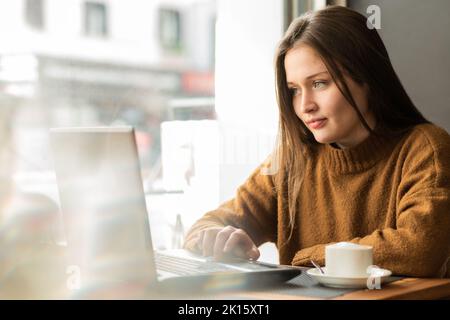 Giovane e premurosa freelancer femminile in caldo maglione con lunghi capelli scuri che guardano via penosamente mentre si siede in un bar con computer portatile e una tazza di cappuccino Foto Stock