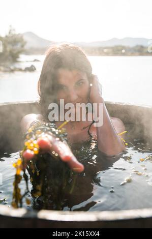 Giovane donna con alghe a contatto con i capelli e raggiungere la macchina fotografica mentre si prende il bagno caldo in barile sulla riva del lago Foto Stock