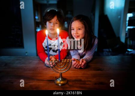 Due bambini si siedono insieme a tavola guardando la menorah illuminata ad Hannukah Foto Stock
